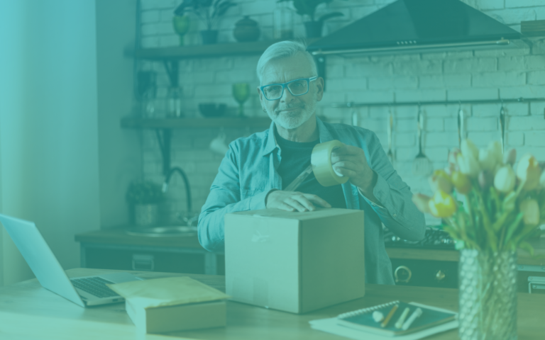 Man wearing glasses is in his kitchen, and he is packaging a box that is sitting on his counter next to flowers and a laptop