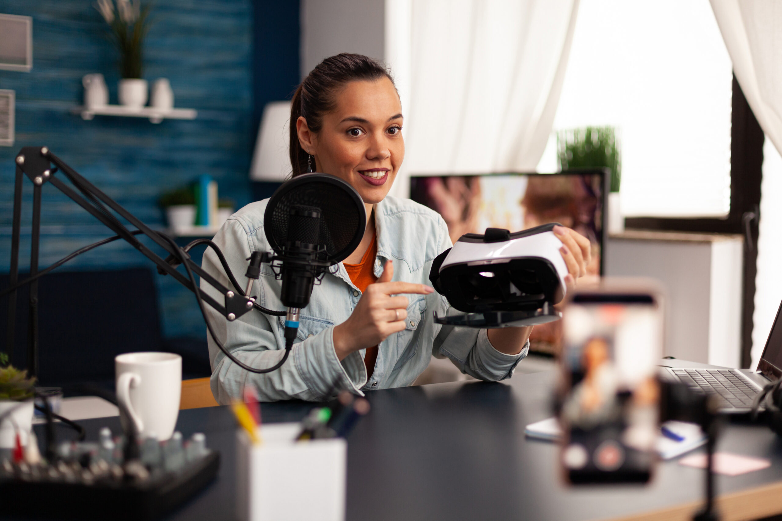 New media influencer smiling at camera while recording vr headphones review for followers. Creative vlogger making video blog concept speaking and looking at smartphone on tripod home studio podcast