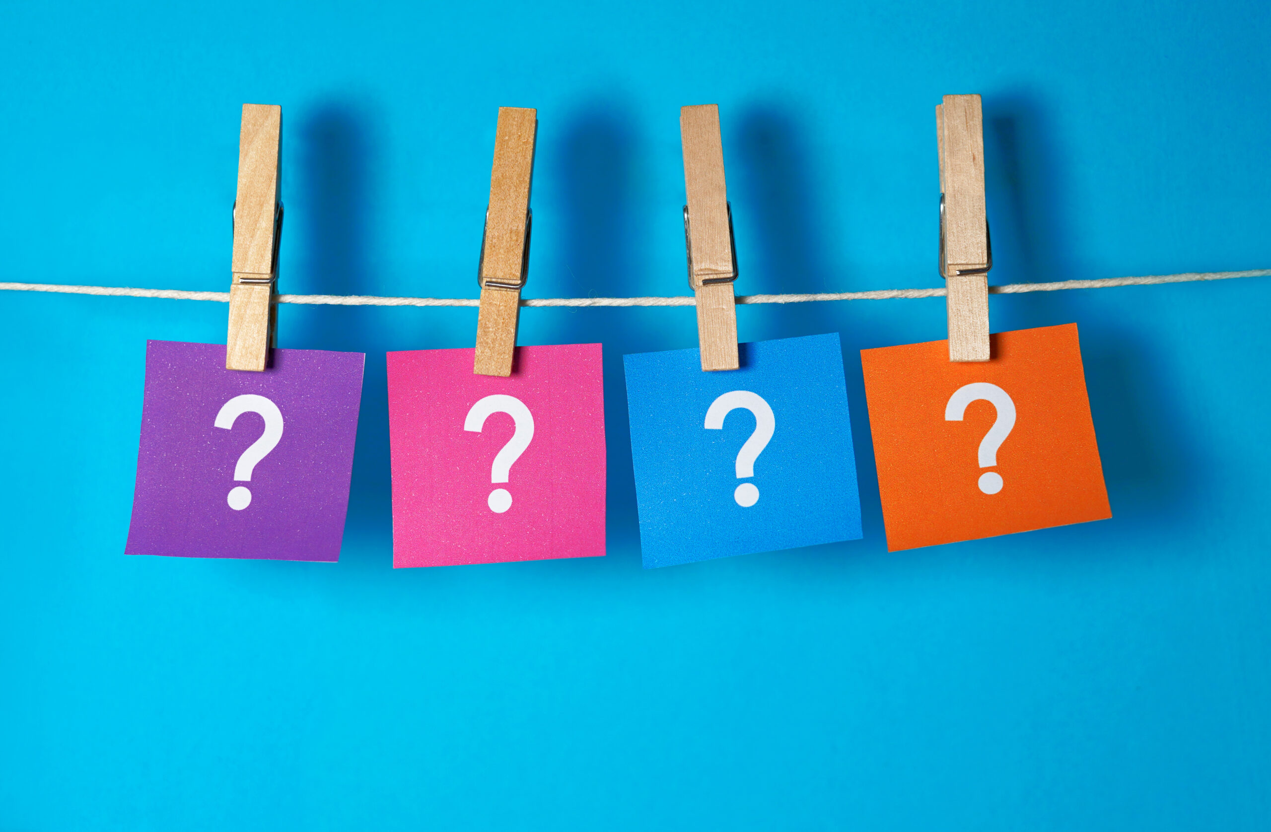Four different colored post-it notes with a question mark on them are clipped to a string using a clothespin.