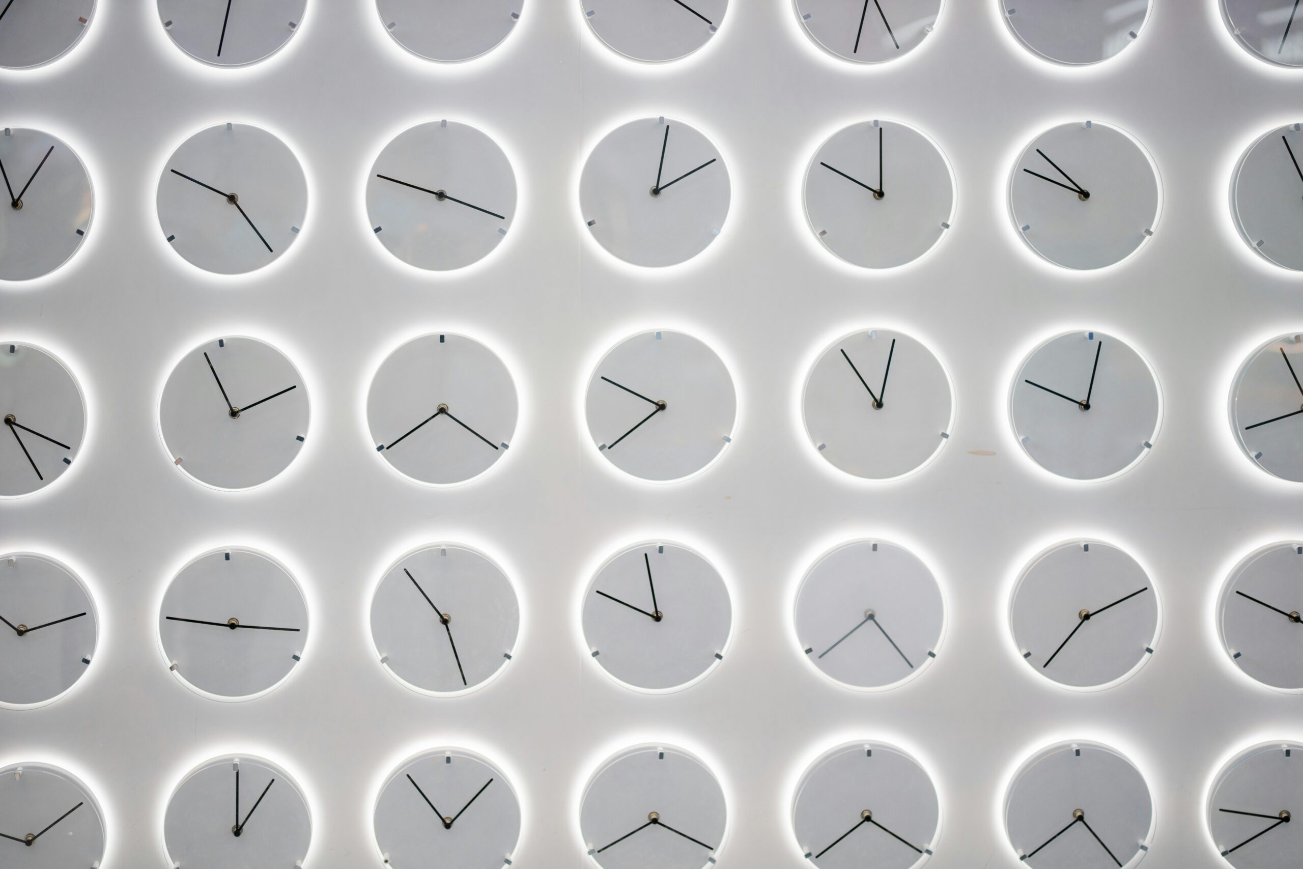 A white wall with a variety of white clocks hung up on it, all showing different times.