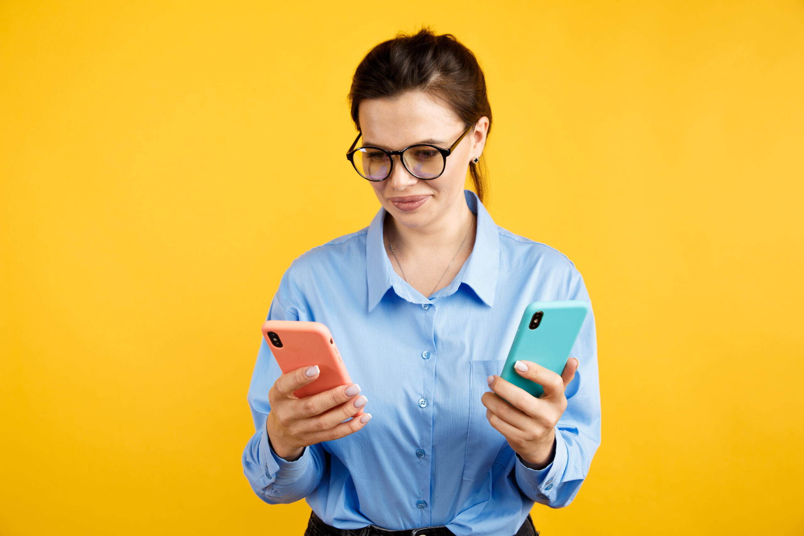 A young professional in front of a yellow background holds one smartphone with a different colored case in each hand.