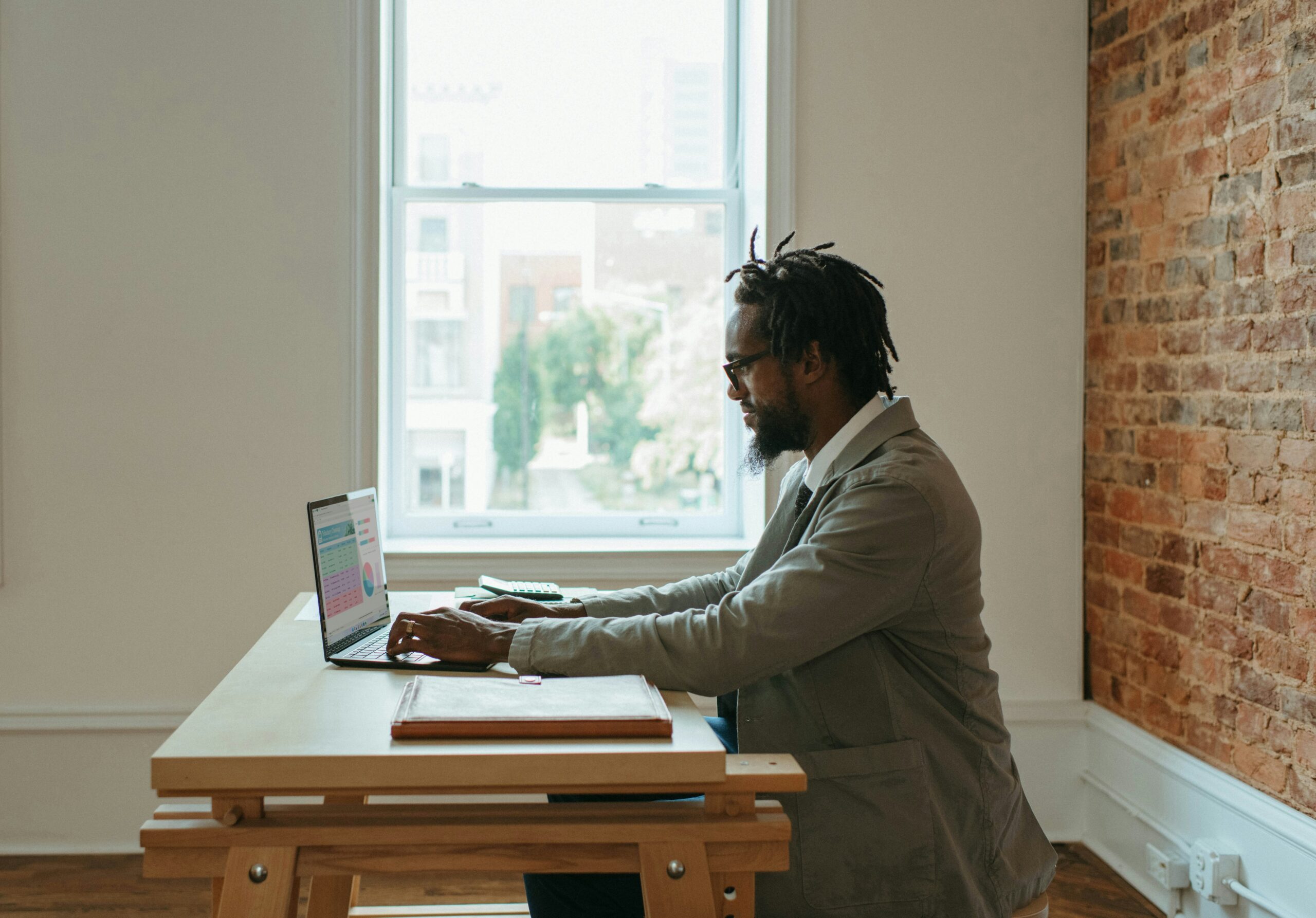 A man sits at a table in front of a window and works on a laptop.