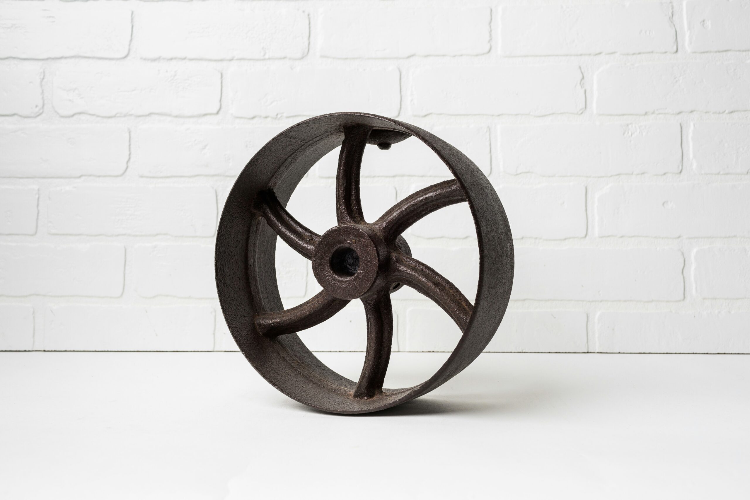 A small brown metal wheel with six spokes sits on a white table in front of a white brick background.