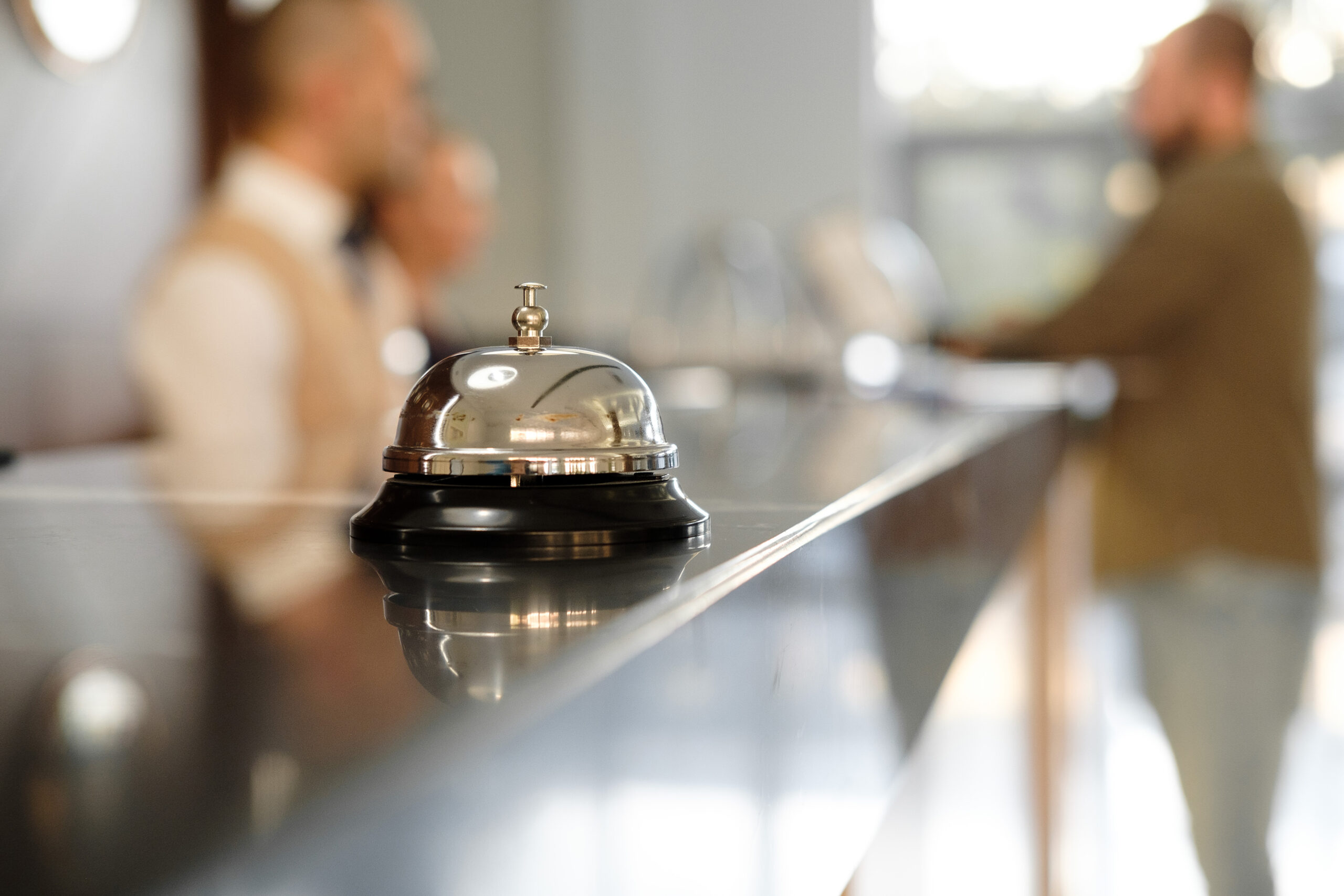 A close up of a bell on a hotel check in counter with three people talking in the background.