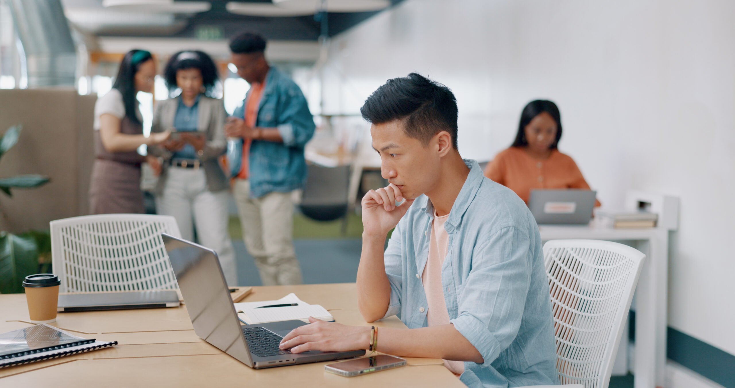 A young professional sits at a desk working on laptop while a group discusses something behind him.